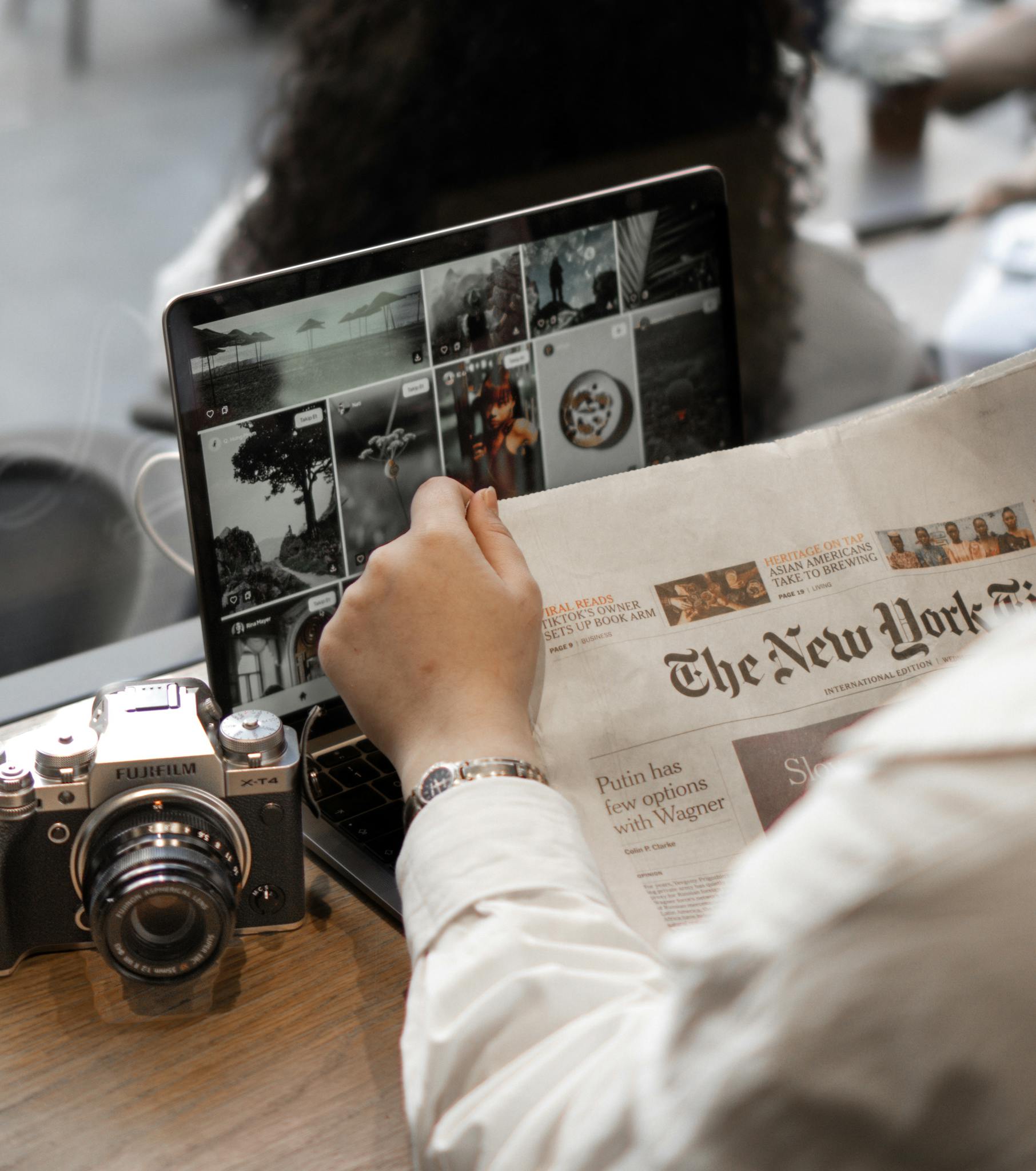 A person multitasks by reading a newspaper and using a laptop, surrounded by a vintage camera in a cafe setting.